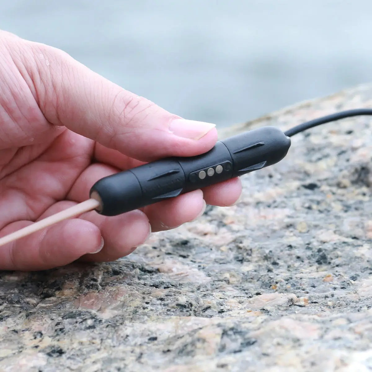 Hand holding a black electronic smart bait with a cable against a blurred natural background